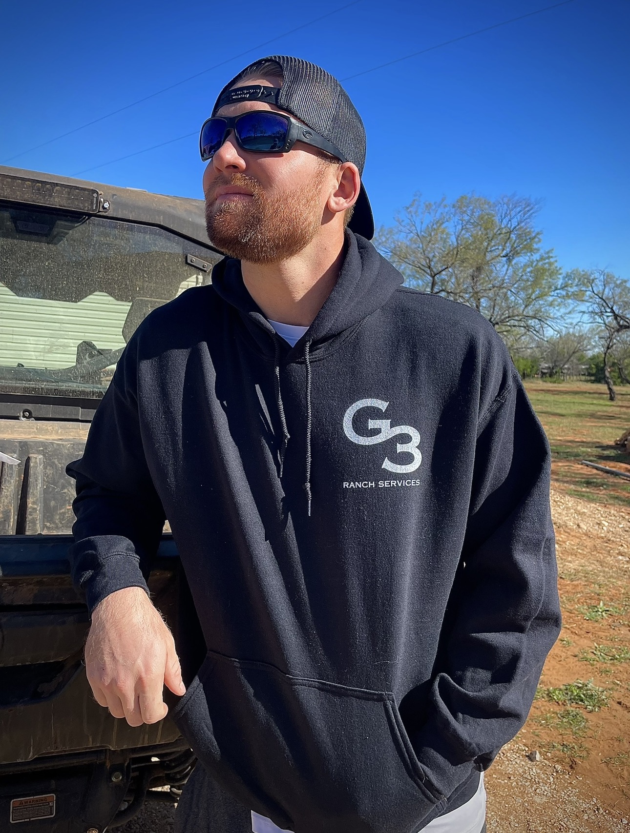 Man wearing a black G3 Ranch Services hoodie and trucker hat leaning against a utility vehicle